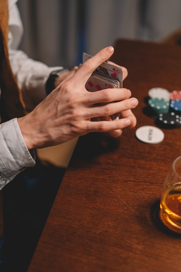 A person shuffling poker cards next to poker chips and a drink on a wooden table.